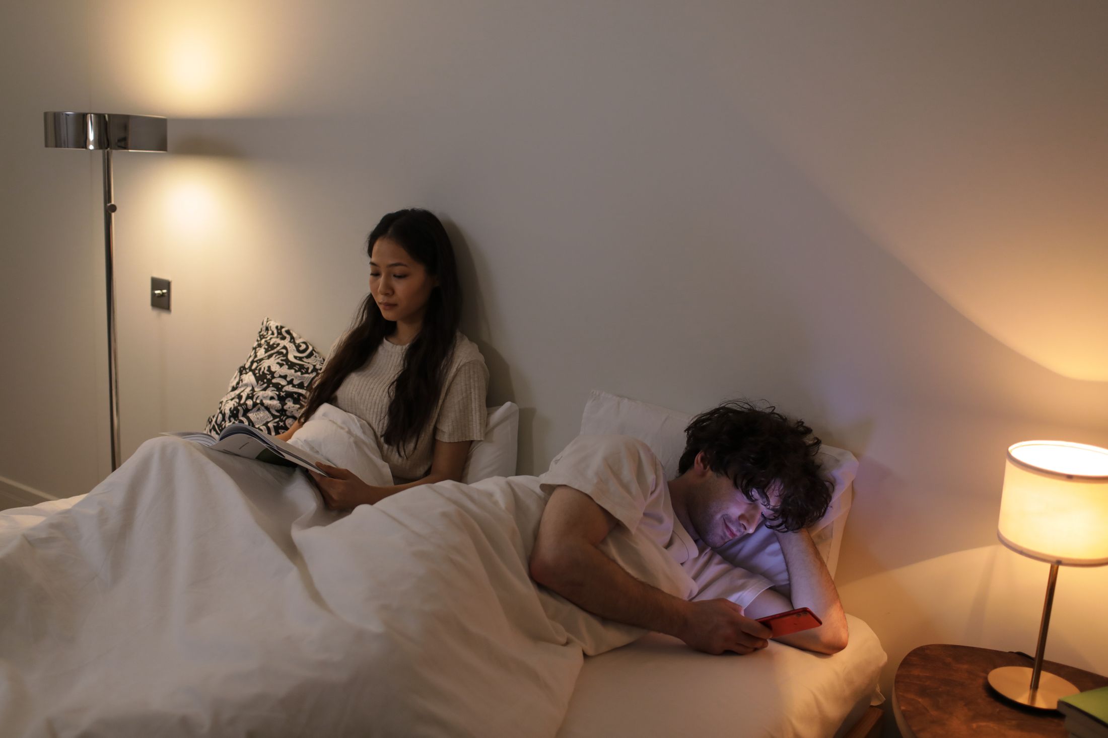 A woman reading in bed beside her husband, who is on his phone